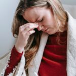Upset serious young female with long blond hair in casual clothes touching forehead with closed eyes while resting in armchair at home