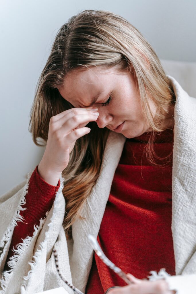 Upset serious young female with long blond hair in casual clothes touching forehead with closed eyes while resting in armchair at home
